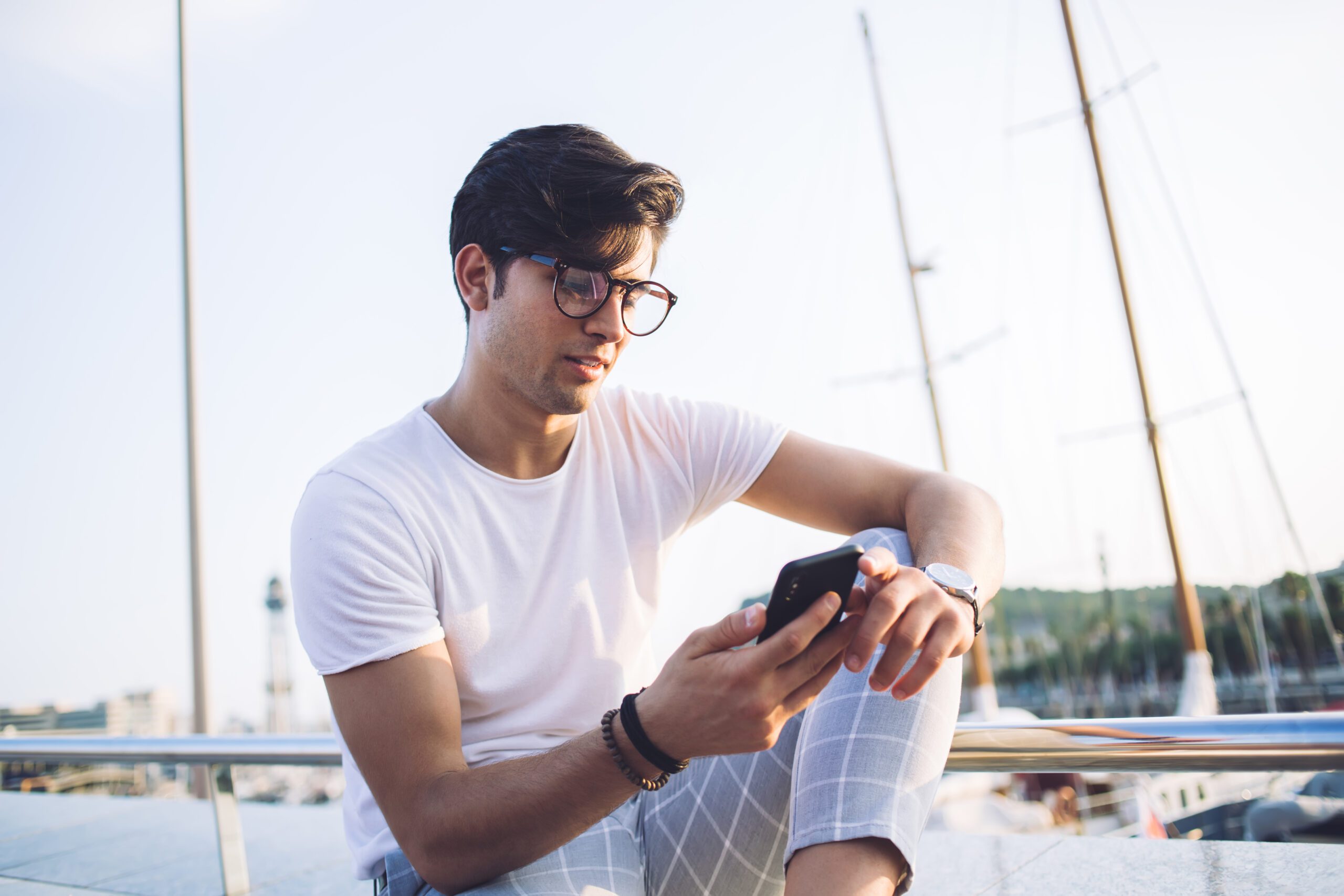 Handsome caucasian male blogger sitting near city harbor share content in blog holding mobile phone and using 4G internet, positive 20s hipster guy read news from social networks near bay in town