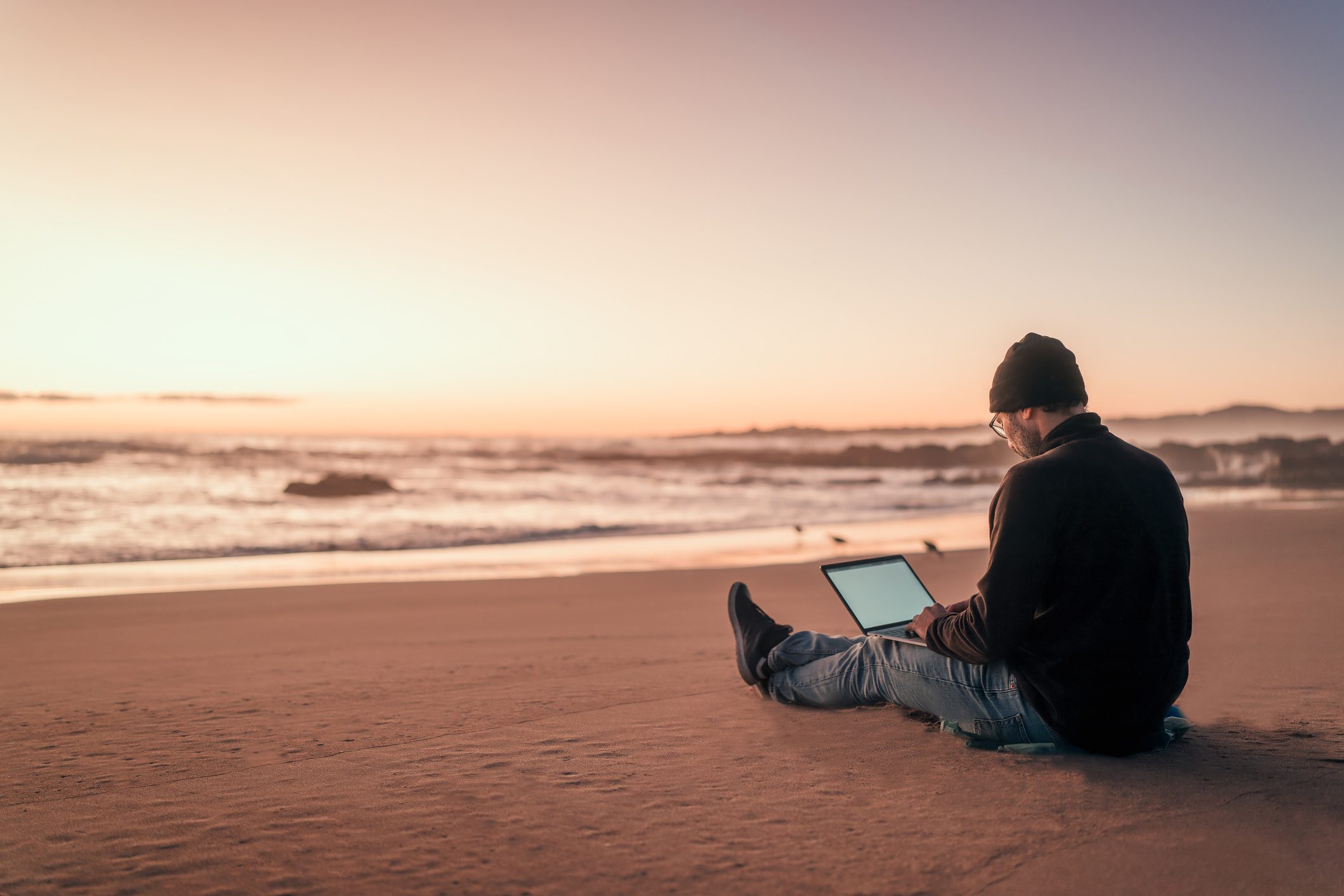 silhouette of a person working on his laptop outdoors on the beach at golden hour, back view